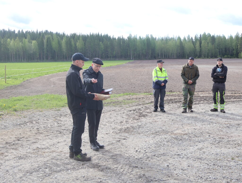 Seurakuntapastori Mikko Elenius siunaa Tommi Harjun pitelemän kylvövakan siemenet. Taustalla toimitusta seuraavat vas. Mikko Viitaniemi, MTK Järviseudun puheenjohtaja Juha Kamppila sekä Harjun lypsytilan kesätyöntekijä Roni Nahkala.