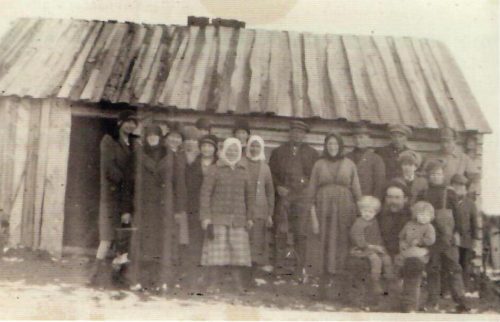 Purmon Kauhajärveläisiä vierailulla Roismassa maaliskuussa 1931.     Perheeseen oli syntynyt pokavauva, ja kyläläiset olivat sen vuoksi               onnittelukäynnillä. Matti istuu kuvan keskellä lapset polvellansa ja Amalia      seisoo hänen vierellään . Mökki oli tuolloin vielä hyvin alkeellinen. Kuva: Anna-Liisa Björklund.